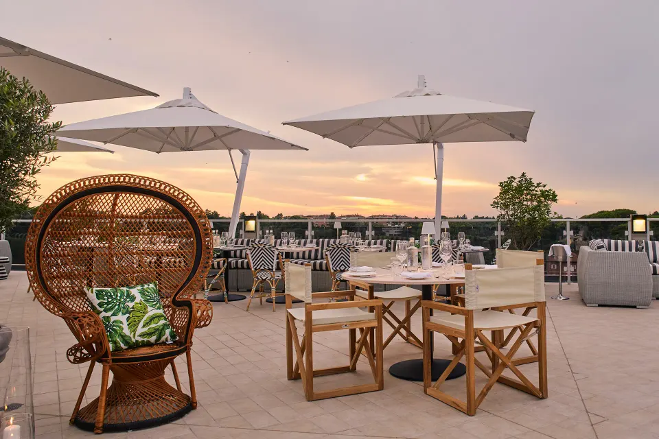 A rooftop terrace at dusk with white parasols, a wicker peacock chair and dining tables overlooking the green park.